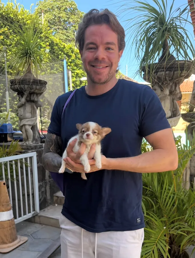 A man holding a fawn and white long haired chihuahua puppy